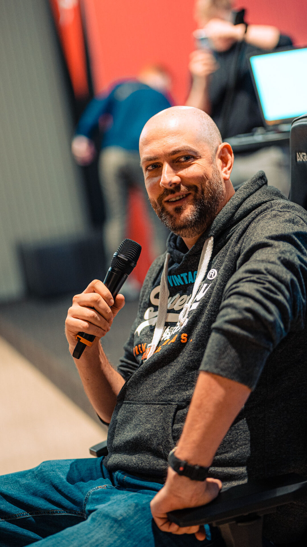 Christian Lenz epf sportlicher Direktor - Er sitzt auf einem Stuhl, schaut seitlich in die Kamera und hält ein Mikrofon in seiner rechten Hand. Im Hintergrund sind unscharf Menschen zu erkennen.