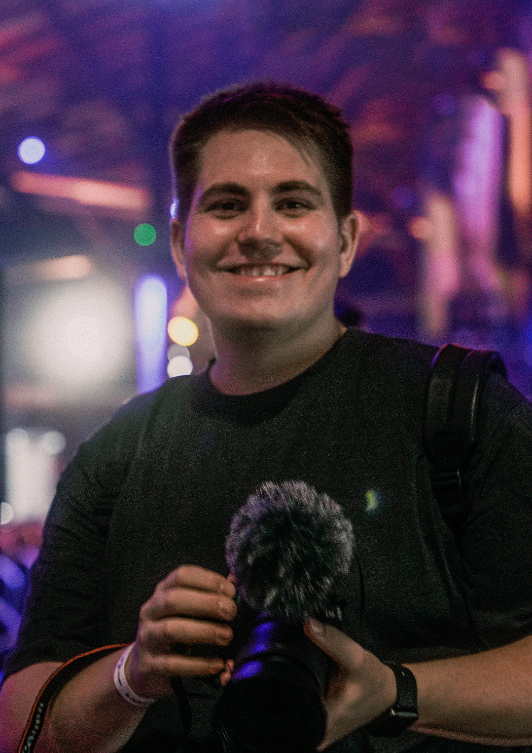 Justin "Pegasus" Wriedt - er lächelt in die Kamera und hält einen Fotoapperat samt Ansteckmikrofon in beiden Händen. Im Hintergrund sind Lichter eines Events zu erkennen.