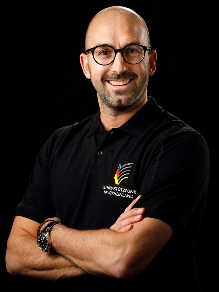 Moritz Anderten Head of Sportspsychology bei der esports player foundation im Portrait - die Arme sind verschränkt und er steht seitlich nach vorne. Auf seinem T-Shirt steht Olympiastützpunkt Deutschland