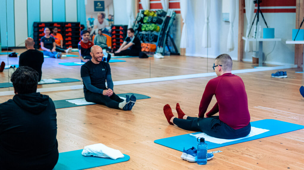 In einem Gym sitzt eine Gruppe von Menschen, die in einem großen Spiegel zu erkennen ist, einer einzelnen Person gegenüber. Diese Person sitzt, die Beine ausgestreckt und spricht zur Gruppe.
