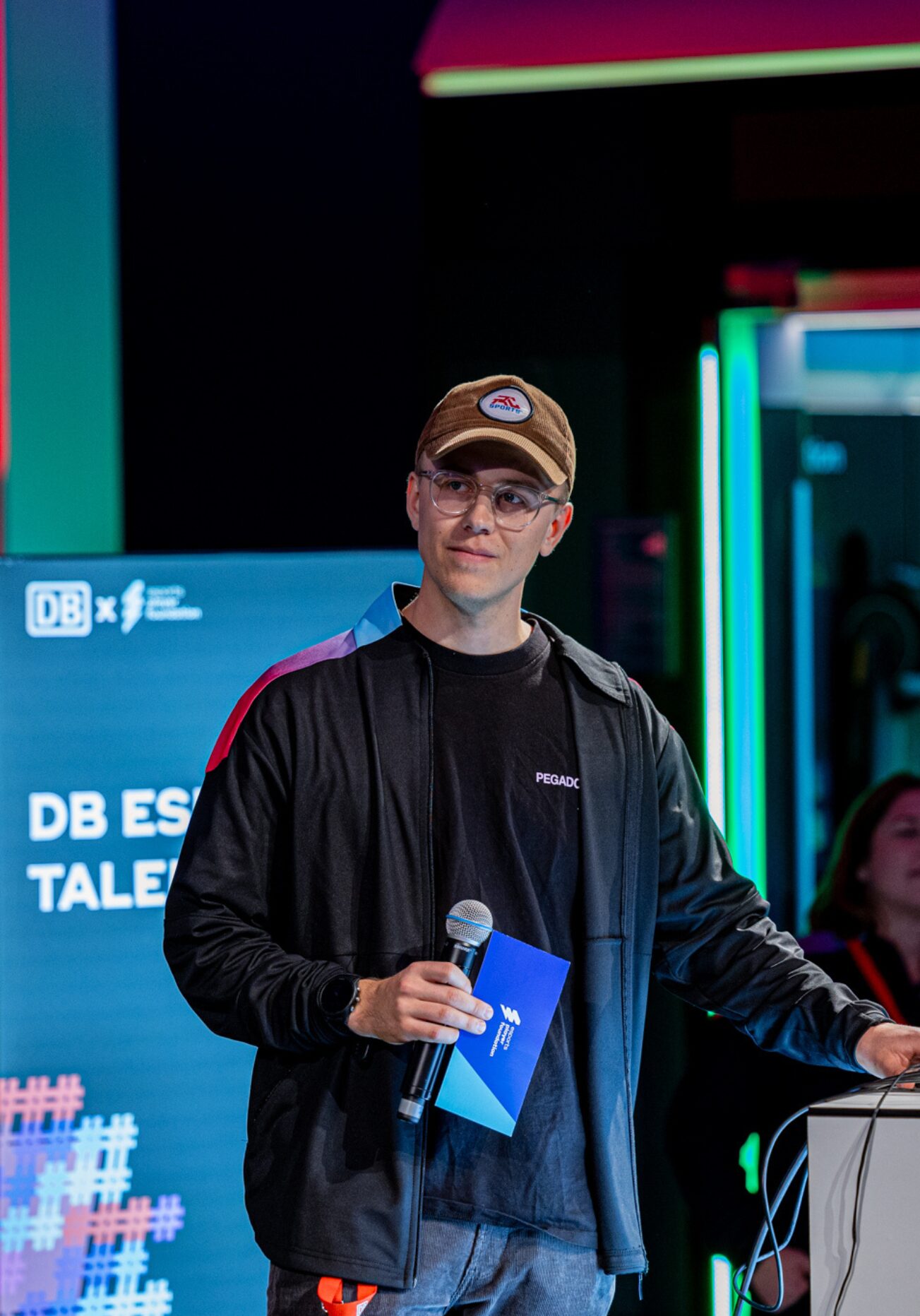 Steffen Wilmers Projektmanager, steht vor einem Podest & hält ein Mikrofon sowie eine Moderationskarte in der Hand