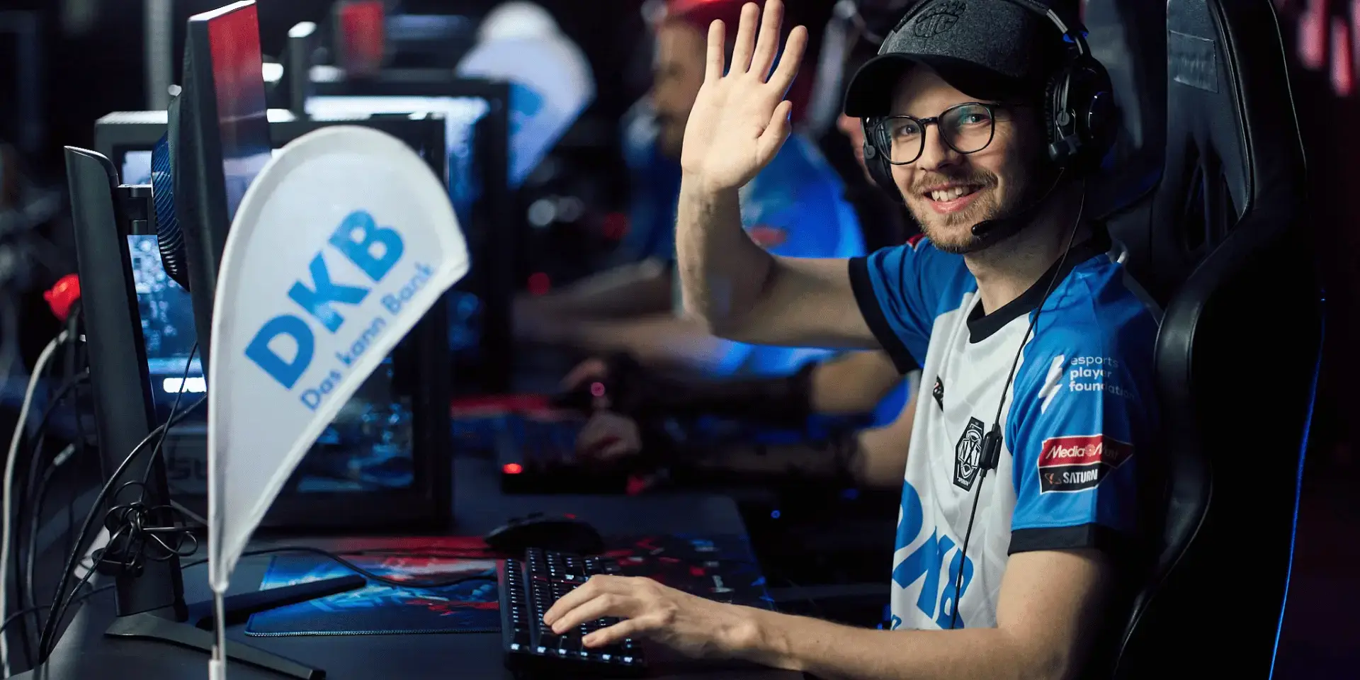 Trainer des DKB XPERION NXT League of Legends Esports Teams sitzt im Trikot vor PC und winkt in die Kamera