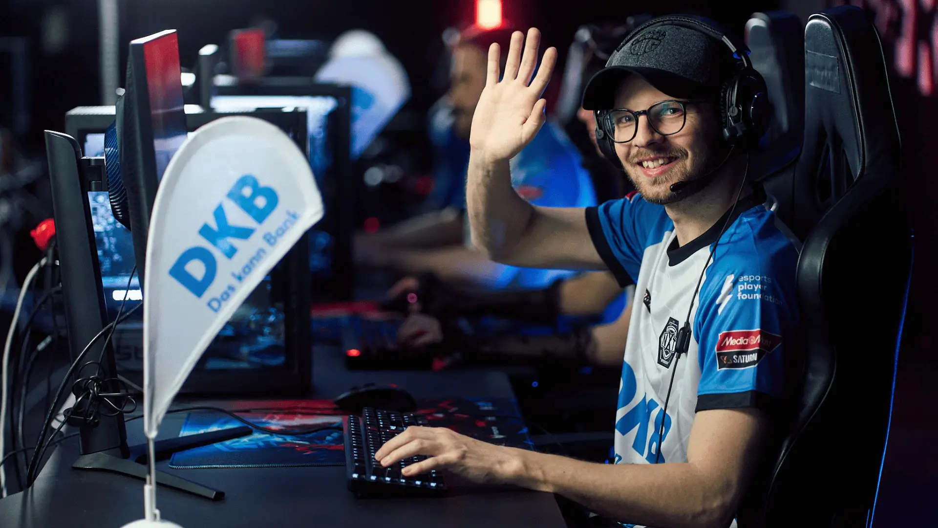 Trainer des DKB XPERION NXT League of Legends Esports Teams sitzt im Trikot vor PC und winkt in die Kamera