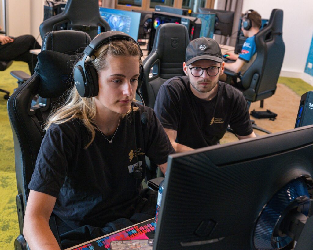 a female player is sitting on a noblechairs gaming chair while being coached in league of legends by a coach sitting next to her
